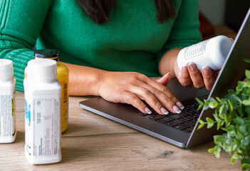 Health and nutrition concept. Indoor image young female holding  food supplement bottle sitting with laptop surfing searching internet for online review of food supplements products on internet.