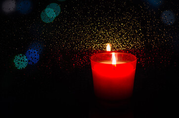 Burning candle in red glass puts beside window with rain drop on bokeh light and dark background.