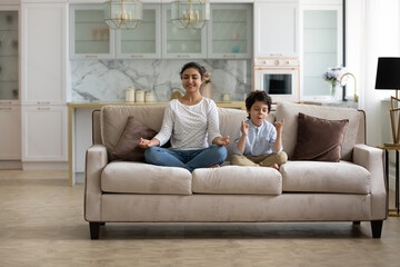 Happy calm Indian mom and little son kid sitting on couch, keeping lotus pose, zen hand gesture, meditating, practicing home yoga, home yoga, mental control, mindfulness, relaxing, exercising