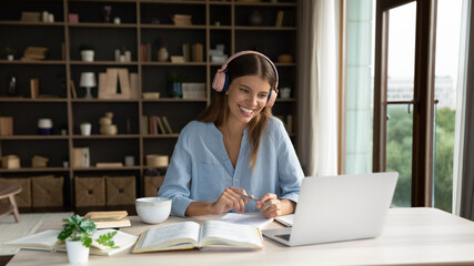 Wall Mural - Happy attractive millennial woman in headphones watching interesting educational online lecture or holding video call with teacher learning foreign language, studying distantly using computer.