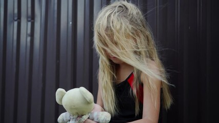 a small blonde girl plays with her doll in front of a metal fence in a refugee camp.