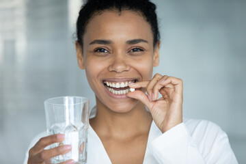 Wall Mural - Close up head shot portrait of beautiful happy young african american mixed race woman taking pills, drinking glass of fresh pure water, daily dose omega vitamins or dietary supplements concept.