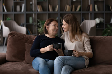 Wall Mural - Happy mature mother with grown-up daughter drinking tea, chatting, sitting on couch at home, smiling young woman and senior mom talking enjoying leisure time, family spending weekend together