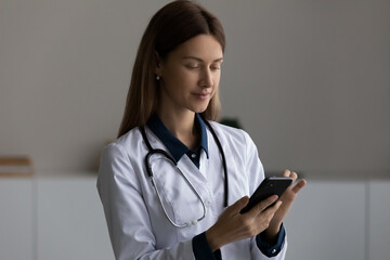 Head shot confident female doctor therapist in white uniform with stethoscope using smartphone, medical apps, looking at modern device screen, woman nurse practitioner consulting patient online