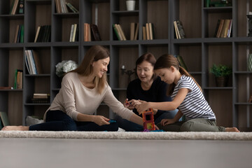 Wall Mural - Little girl with mother and grandmother having fun, sitting on warm floor at home, three generations of women spending leisure time together, playing with toys colorful plastic blocks, building tower