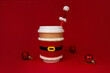 © ksenija1803z - paper beige cup with white lid dressed in santa claus clothes, red Christmas hat and balls on scarlet background