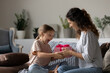© fizkes - Loving smiling mother giving pink gift box to surprised little daughter, congratulating with birthday, sitting on couch, overjoyed cute girl child excited by present, family celebration concept