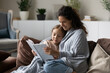 © fizkes - Smiling caring mother with little daughter reading book together, happy mom with adorable girl child wrapped in warm blanket sitting relaxing on comfortable couch, engaged in educational activity