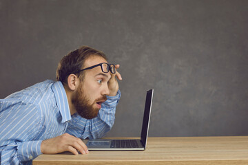 Side view of a man with a surprised expression sitting at a table and staring at a laptop screen. Adult man raises his glasses looking at shocking content or surprised by software bug.