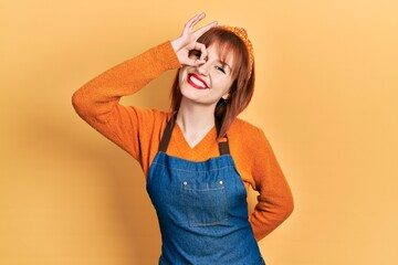 Wall Mural - Redhead young woman wearing apron smiling happy doing ok sign with hand on eye looking through fingers