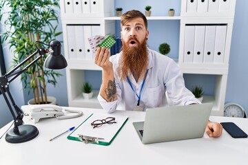 Poster - Redhead man with long beard wearing doctor uniform holding prescription pills scared and amazed with open mouth for surprise, disbelief face