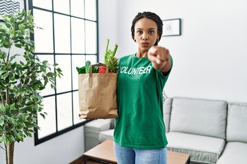 Wall Mural - Young african american woman wearing volunteer t shirt holding bag of groceries pointing with finger to the camera and to you, confident gesture looking serious