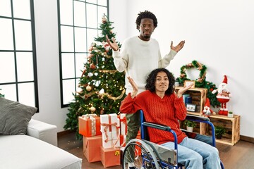 Poster - Young interracial couple with woman sitting on wheelchair by christmas tree clueless and confused expression with arms and hands raised. doubt concept.