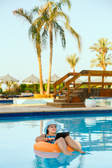 Wall Mural - A Jewish woman in a sun hat in the pool in a swimming circle with a cocktail and a laptop on a background of palm trees.