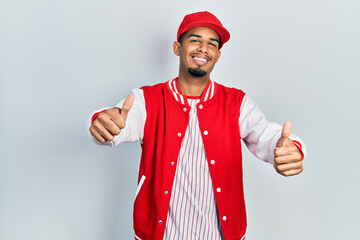 Young african american man wearing baseball uniform approving doing positive gesture with hand, thumbs up smiling and happy for success. winner gesture.