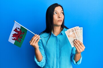 Poster - Young latin woman holding wales flag and pounds banknotes smiling looking to the side and staring away thinking.