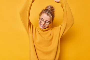 Wall Mural - Positive carefree woman raises arms up dances carefree from joy wears round transparent glasses and jumper smiles gladfully keeps eyes closed isolated over vivid yellow background rejoices success