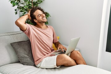 Poster - Young hispanic man sitting on the sofa at home using laptop smiling confident touching hair with hand up gesture, posing attractive and fashionable