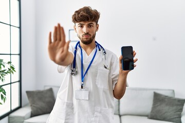 Sticker - Young arab man wearing doctor uniform and stethoscope holding smartphone with open hand doing stop sign with serious and confident expression, defense gesture