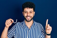 Young Arab Man With Beard Holding Invisible Aligner Orthodontic And Braces Smiling With An Idea Or Question Pointing Finger With Happy Face, Number One
