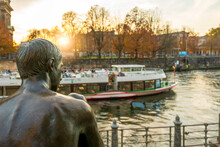 Bronze Boy Statue Overlooking Spree River In Berlin While A Tourist Boat Is Passing By At Sunset Of An Autumn Day.  Berlin, Germany