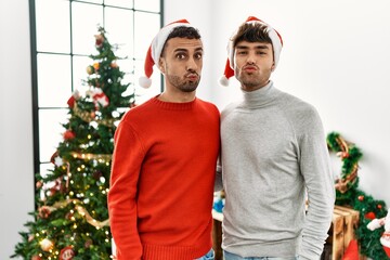 Poster - Young gay couple standing by christmas tree wearing hat looking at the camera blowing a kiss on air being lovely and sexy. love expression.