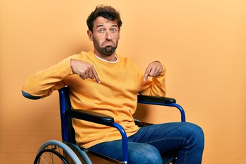 Sticker - Handsome man with beard sitting on wheelchair pointing down looking sad and upset, indicating direction with fingers, unhappy and depressed.