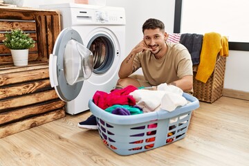 Wall Mural - Young handsome man putting dirty laundry into washing machine smiling doing phone gesture with hand and fingers like talking on the telephone. communicating concepts.