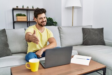 Poster - Young man with beard using laptop at home smiling friendly offering handshake as greeting and welcoming. successful business.