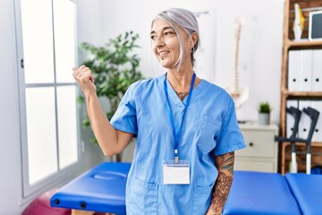 Poster - Middle age grey-haired woman wearing physiotherapist uniform at medical clinic smiling with happy face looking and pointing to the side with thumb up.