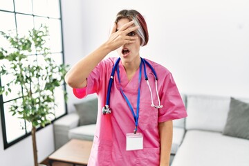 Canvas Print - Young beautiful woman wearing doctor uniform and stethoscope peeking in shock covering face and eyes with hand, looking through fingers with embarrassed expression.