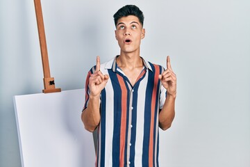 Canvas Print - Young hispanic man standing close to empty canvas amazed and surprised looking up and pointing with fingers and raised arms.