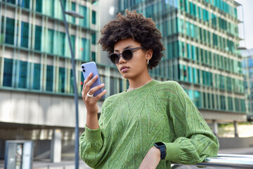 Wall Mural - Fashionable woman chats with friends in social networks surfs on internet website for searching interesting places in city holds cellular wears sunglasses and casual jumper poses near office building