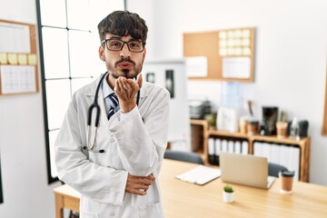 Poster - Hispanic man with beard wearing doctor uniform and stethoscope at the office looking at the camera blowing a kiss with hand on air being lovely and sexy. love expression.