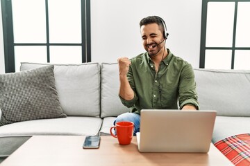 Wall Mural - Young hispanic man with beard wearing call center agent headset working from home very happy and excited doing winner gesture with arms raised, smiling and screaming for success. celebration concept.