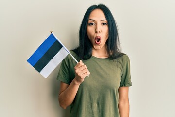 Wall Mural - Young hispanic girl holding estonia flag scared and amazed with open mouth for surprise, disbelief face