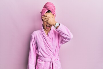 Wall Mural - Young blonde woman wearing shower towel cap and bathrobe smiling and laughing with hand on face covering eyes for surprise. blind concept.