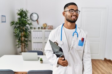 Poster - Young indian man wearing doctor uniform and stethoscope looking away to side with smile on face, natural expression. laughing confident.