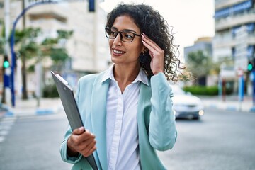 Wall Mural - Young hispanic business woman wearing professional look smiling confident at the city holding worker clipboard