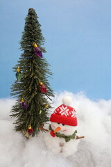 Wall Mural - Evergreen with Colored Bulbs and Snowman With Red Knit Cap on Cottony Snow with Blue Background