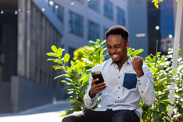 happy african american man in casual clothes sitting on bench outdoors with mobile phone in hand reading good news, winning victory looking at smartphone rejoices outside. Guy emotions student result