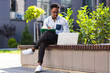 © Liubomir - Young african american male student sitting in a city park on a bench with laptop and notebook studying online outdoors Man freelancer in glasses with books learn working remotely in street e-learning