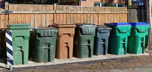 Trash and recycling cans lined up against wooden fence in neighborhood alley.