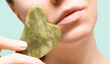 Female model hands hold green jade gua sha scraper in front of her face. Spa body care tool. Beauty care gua sha stone scraper. Copy space.