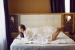 © saulich84 - woman in a white bathrobe is sitting on the edge of the bed and drinking tea from mug at a large window in the hotel