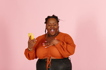 Excited young black plus size body positive woman with dreadlocks in orange top holds mobile phone looking at camera on light pink background in studio closeup