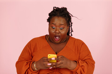 Wondered young black plus size body positive woman with dreadlocks in orange top holds smartphone standing on light pink background in studio closeup