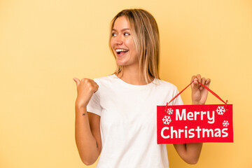 Wall Mural - Young caucasian woman holding a merry christmas placard isolated on yellow background points with thumb finger away, laughing and carefree.