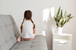 © sementsova321 - Back side of little child sitting on gray sofa, kid wearing white casual t shirt, dark haired female child with pigtail posing backwards to camera, feeling sorrow and sadness.
