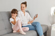 © sementsova321 - Indoor shot of attractive Caucasian woman with hair bun sitting on sofa and talking via video call, her shy daughter does not want to see at cell phone camera.